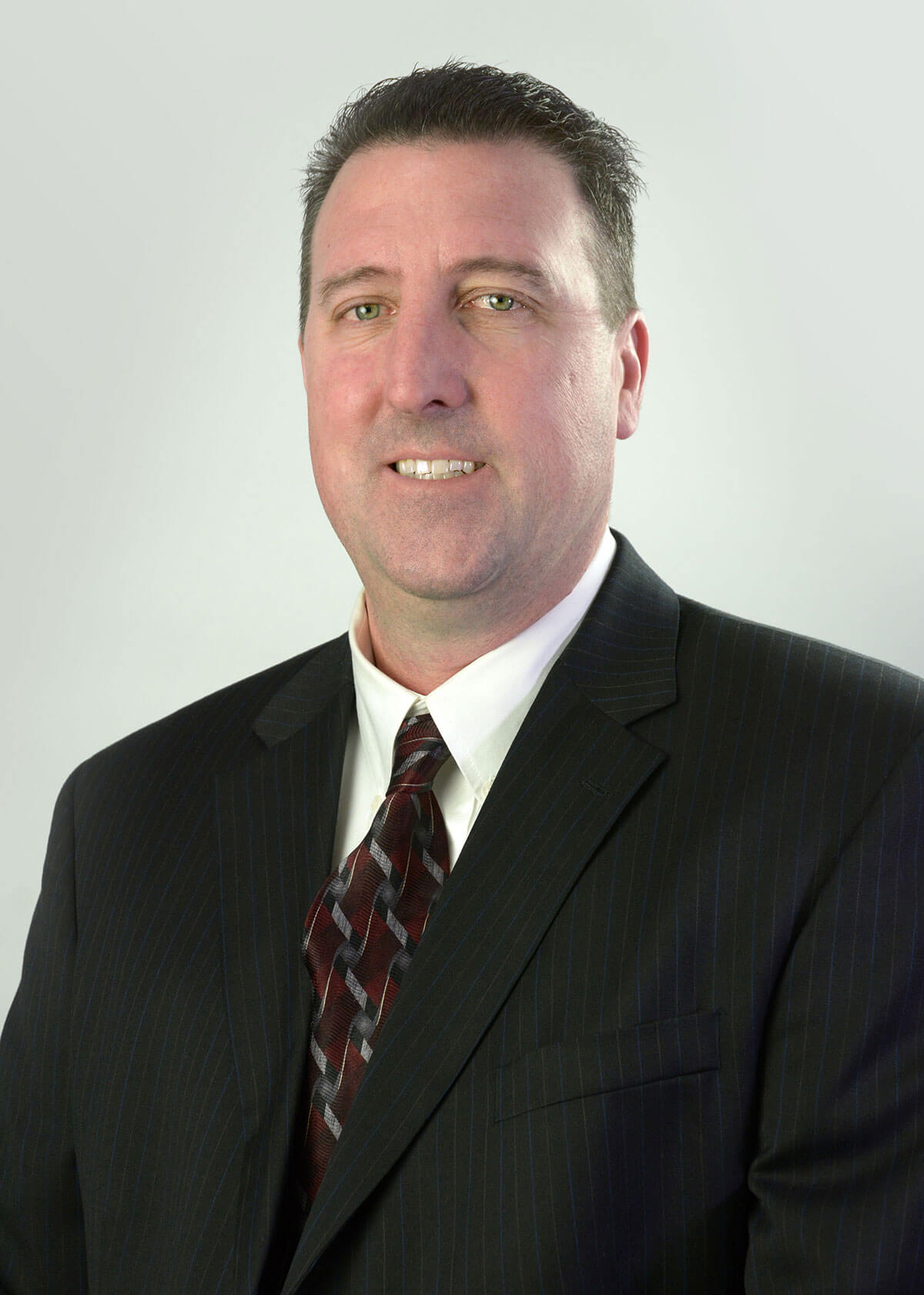 Eric Schwertley smiling in front of a light gray backdrop wearing a black suit coat, a white button up shirt, and a red, black, and gray tie