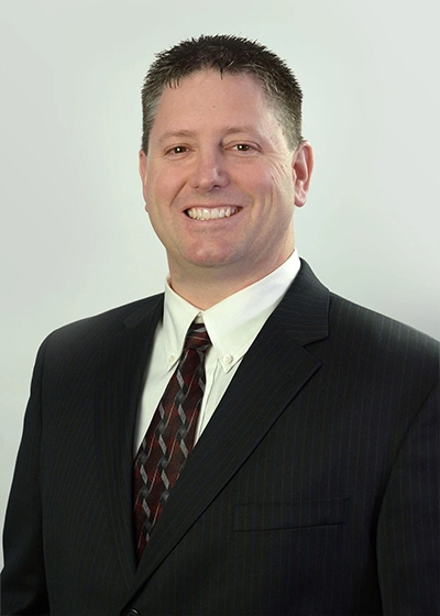 Pete Simonson smiling from the chest up while wearing a black pinstrip suit coat, a white button up shirt, and a red tie with black and white designs on it